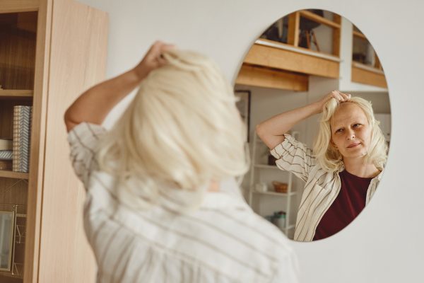 Warm-toned back view portrait of bald woman taking off wig while looking in mirror in home interior, alopecia and cancer awareness, copy space